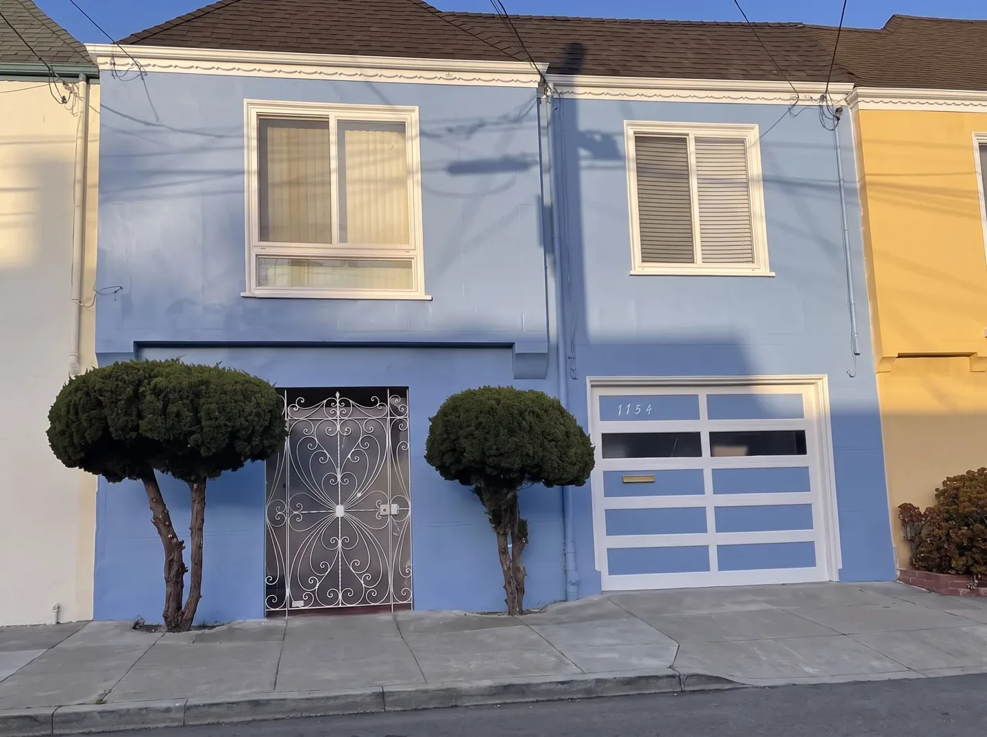 Two-storey house repainted light blue with white trim and a white garage door, two round shrubs by the gate